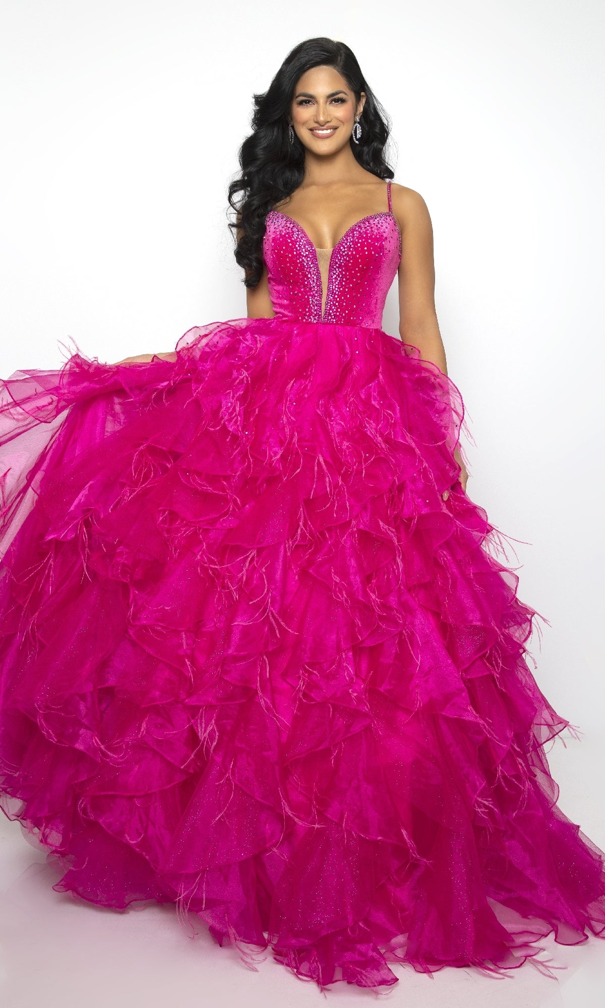 A woman smiles at the camera in the Ava Presley 39554, a bright pink long ruffled prom ball gown with a deep V-neckline and beaded bodice, standing against a plain white background.